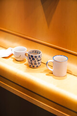 Three ceramic mugs of different designs and colors sit on a wooden surface in warm lighting. A cozy and nostalgic atmosphere, perfect for themes of home, coffee, tea, relaxation, and daily rituals.