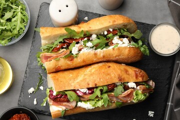 Halves of baguette sandwich with prosciutto and sun dried tomatoes and ingredients on grey table, flat lay