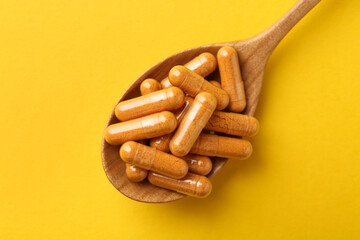 Turmeric capsules in spoon on yellow background, top view