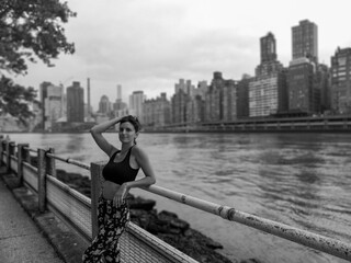 Black and white. A lady stands by a lamppost, gazing at the New York skyline, encapsulating the city's beauty and allure in a timeless black and white photograph.
