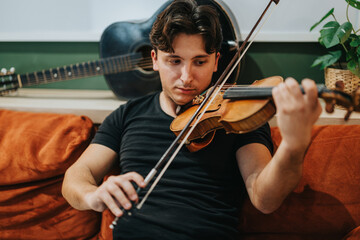 A young man seated on a comfortable couch, attentively playing a wooden violin. Behind him, a...