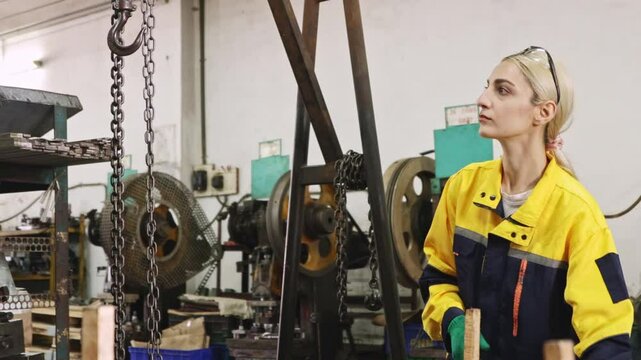 Female factory worker wearing safety glasses and gloves pulling chain on steel beam for safety while lifting and moving heavy machinery placed on floor in automotive parts lathe factory.