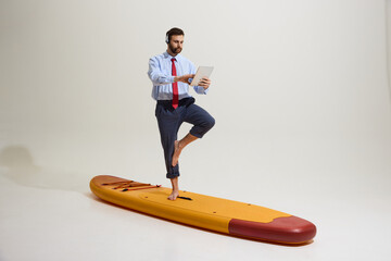 Businessman in rolled-up formal pants, tie, and headset balancing on one leg on paddleboard,...