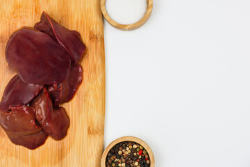 Close-up on the left on raw chicken liver on a wooden board, next to wooden bowls of salt and pepper seasoning on a white background with space for text. The concept of healthy and healthy eating