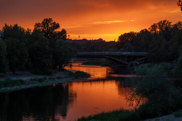 Fototapeta premium Stunning sunset over a calm river with a silhouette of a bridge and trees at twilight