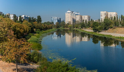 Fototapeta premium Lush riverside park in an urban setting with reflections of buildings on a clear day. Kyiv city 