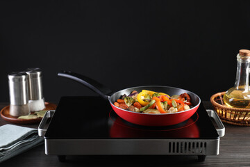 Frying pan with vegetables and stove on wooden table against black background