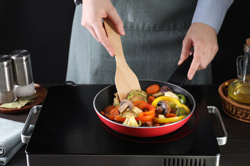 Woman frying vegetables and mushrooms in pan on stove at wooden table, closeup