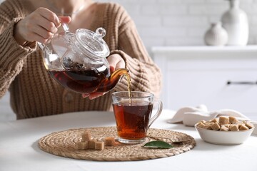 Woman pouring hot black tea into cup at white table, closeup