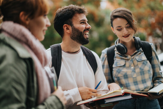A group of young students discussing in an outdoor setting, holding books and wearing backpacks. They appear engaged and enthusiastic about their studies. The mood is collaborative and friendly.