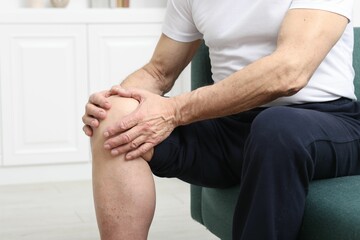 Senior man suffering from pain in his knee on sofa indoors, closeup