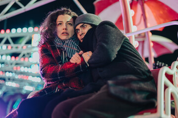 Fototapeta premium A worried couple sits closely together at a fairground while vibrant lights illuminate the scene. The fairground atmosphere contrasts with their serious expressions.
