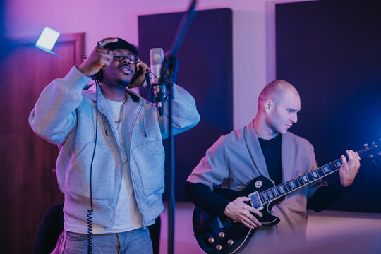 Two musicians work together in a recording studio, one singing into a microphone and the other playing an electric guitar. The setting has vibrant lighting and a modern, creative atmosphere.