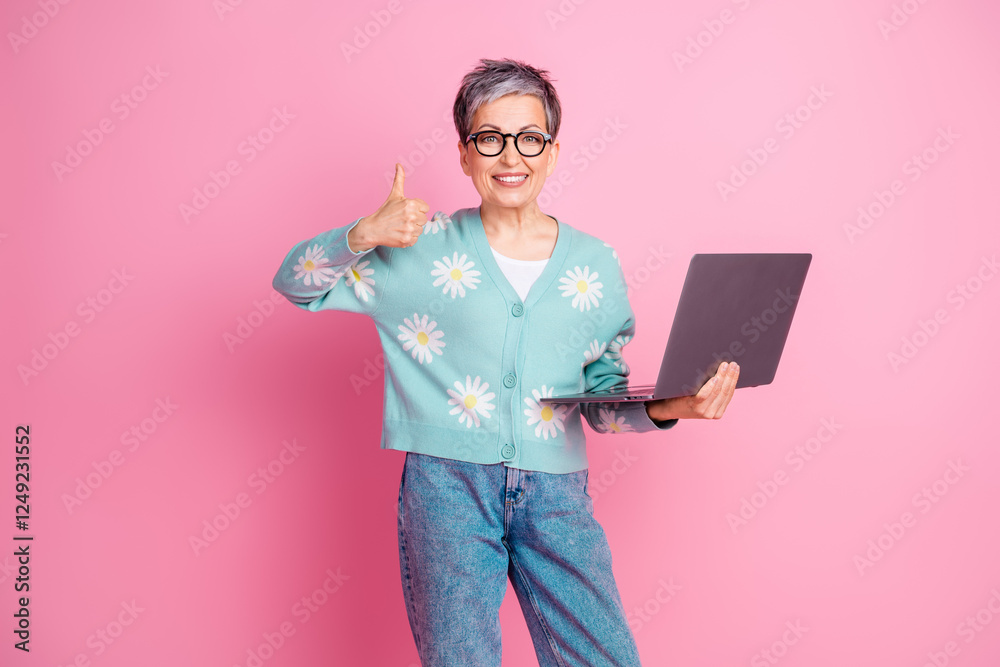 Wall mural Happy mature woman holding laptop and showing thumbs-up gesture on pink background