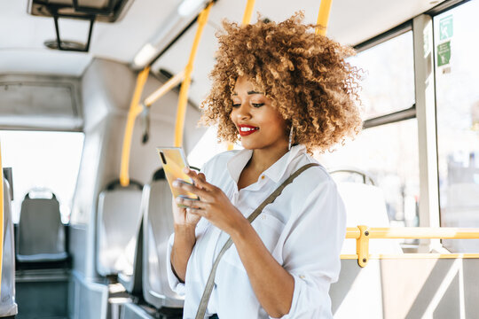 Beautiful and fashionable black woman standing in city bus. She is happy and smiled while using her smartphone to communicate with someone. Modern city lifestyle concept. Bright sunny day.