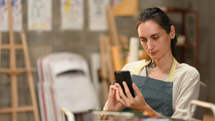 An artist in an apron focuses on her smartphone while seated in a creative studio filled with easels, sketches, and art supplies, balancing art and modern technology seamlessly
