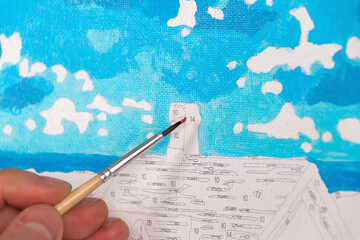 A hand painting a numbered canvas, depicting a bright blue sky with white clouds. Painting by...