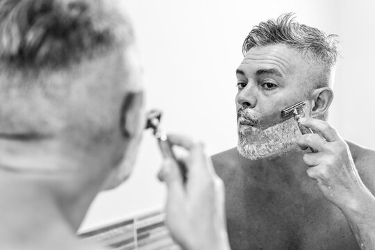Man shaving with a safety razor in front of a bathroom mirror. The black and white composition emphasizes the grooming routine, self-care, and traditional wet shaving practices.