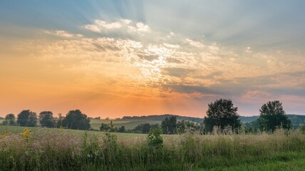 Obraz premium A serene landscape at sunset, showcasing vibrant orange and blue hues with sun rays breaking through fluffy clouds over lush fields and trees.