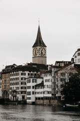 Fototapeta premium Iconic clock tower rising above traditional European buildings, with a calm river in the foreground under an overcast sky.