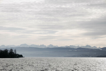 Wide view of a calm lake with mountain ranges in the distance under a cloudy, overcast sky.