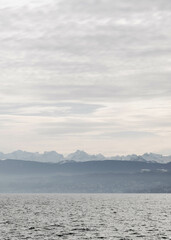 Wide view of a calm lake with mountain ranges in the distance under a cloudy, overcast sky.