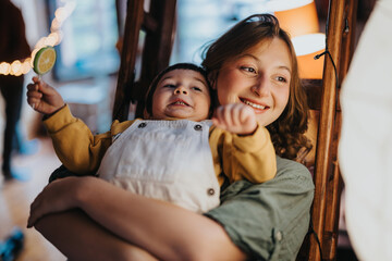 Obraz premium Smiling woman holding a young child, they both appear cheerful. The bright and lively indoor atmosphere conveys happiness and warmth, capturing a moment of family connection.