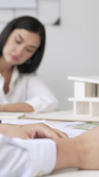 Professional architect engineer drawing blueprint and working together with young beautiful caucasian coworker on meeting table with safety helmet, house model, blueprint scatter around. Immaculate.