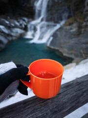 Gloved hand holding steaming orange mug near cascading waterfall, misty november landscape in aosta...