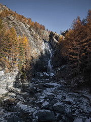 Cascading waterfall dropping between rocky cliffs, saturated autumn colors enveloping landscape near aosta, italian mountainous terrain Cascate di Lillaz