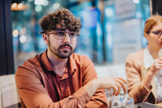 A young man attentively listens during a business meeting, seated at a modern office table. The atmosphere is professional, with a blurred background indicating a busy office setting. - Powered by Adobe