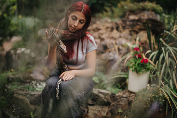 A girl sits in a lush garden, calmly holding a snake. The natural surroundings and her serene...
