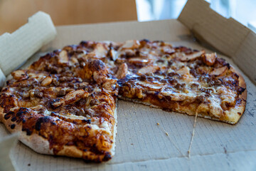 Close-up of a freshly baked pizza in an open takeaway cardboard box with one slice missing. Topped with barbecue sauce, chicken, beef, and bacon. Perfect for food delivery themes