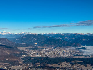 The city of Villach lies nestled in alpine valley, viewed from the Mallestiger Mittagskogel. Snow-capped mountain peaks rise in the distance under a blue sky, creating a scenic Austrian landscape.