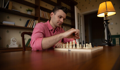 A man in a pink shirt thoughtfully moves a chess piece on a wooden chessboard. The room has bookshelves, a clock, and a lamp, creating a cozy and focused atmosphere for the game.
