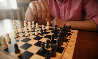 A person in a pink shirt moves a white chess piece on a wooden chessboard. The game is in progress, with black and white pieces thoughtfully placed on the board, set in a cozy room.