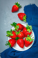 Fresh strawberries on a white plate and a gray textured background, complemented by a dark blue fabric. Bright contrast of colors, top view. Flat lay.