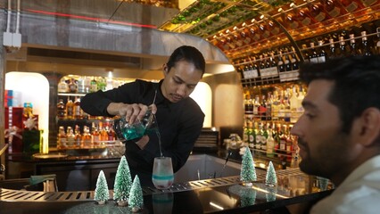 Bartender pouring mixed blue lagoon cocktail liqueur to cooling glass while guest talking friends waiting drink, decorated pine light christmas theme making special festive tasty in couple. Vinosity.