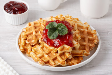 Delicious waffles topped with strawberry jam and a fresh leaf on a white plate with a light wooden background