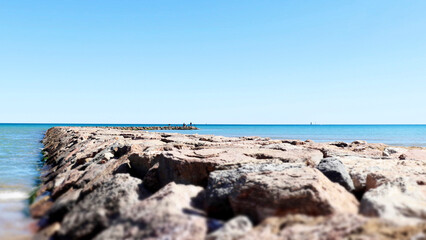 A stone breakwater extending into the azure sea under a bright blue sky. The warm sunny day, clear horizon, and endless waters evoke a sense of tranquility and freedom.