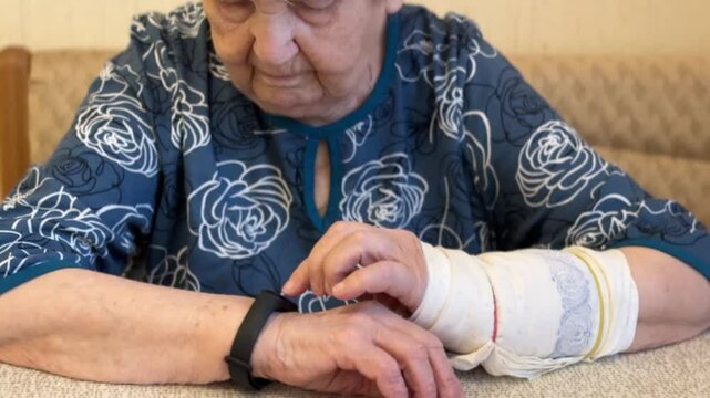 The black smart band with display touchpad and heart rate monitor. The old woman with broken arm with cast for the arm sitting at sofa touches and looks at the wristband of pulse monitor indoors