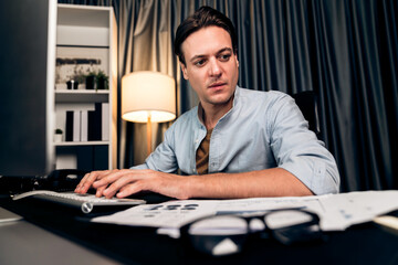 Obraz premium Smart businessman typing with keyboard on monitor with business paragraph dynamic data marketing analysis planning sheet aligning on report paperwork at modern home office at night time. Pecuniary.