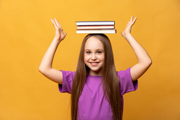 Happy school child girl in lilac t-shirt with books on head and hands raised up on yellow...