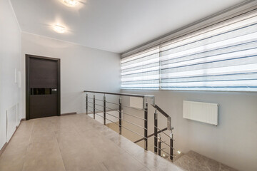 A hall with a black door and a staircase in a private house. The wide window is covered with curtains.