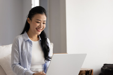 Positive online project manager working at laptop from home. Happy beautiful young Asian freelancer...