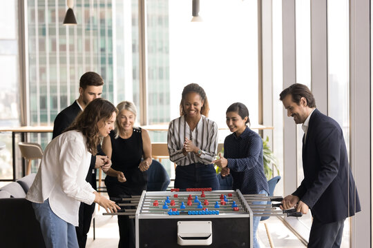 Group of multiracial colleagues gathered around foosball table in modern, high-rise office, engaged in playing game of tabletop soccer in lively and friendly atmosphere. Fun, lunch break at workplace