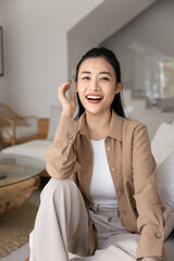 Cheerful beautiful young Asian homeowner woman resting on cozy home couch, touching facial skin,...