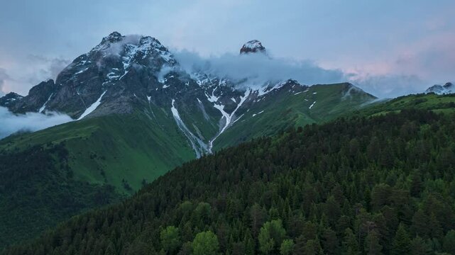Aerial hyperlapse of Ushba peak at sunset in Caucasus mountains, Svaneti. Majestic mountains, snow-capped peaks, clouds, green forest. Nature, travel exploration, adventure video Drone shot