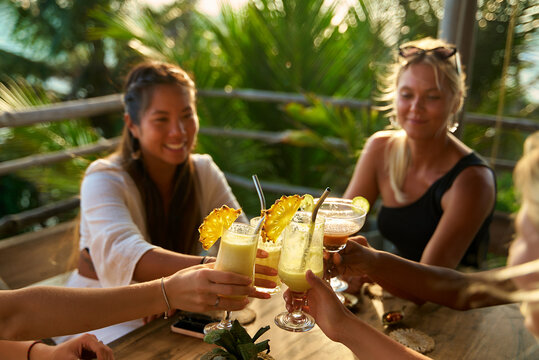 Diverse group of women at tropical restaurant toast with cocktails. Celebration. Vacation mood. Fun bachelorette party on travel. Laughter in paradise. Enjoying sun vibes together. Happy moments.