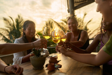 Diverse women cheers with wine glasses at sunset restaurant. Friends toast, enjoy drink, social meetup. Wine tasting, women club. Celebrate friendship. Relax, chat, smile outdoors. Sunset.